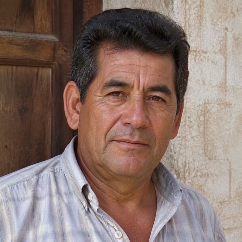 Uomo maturo con capelli scuri e camicia a righe, sfondo rustico.